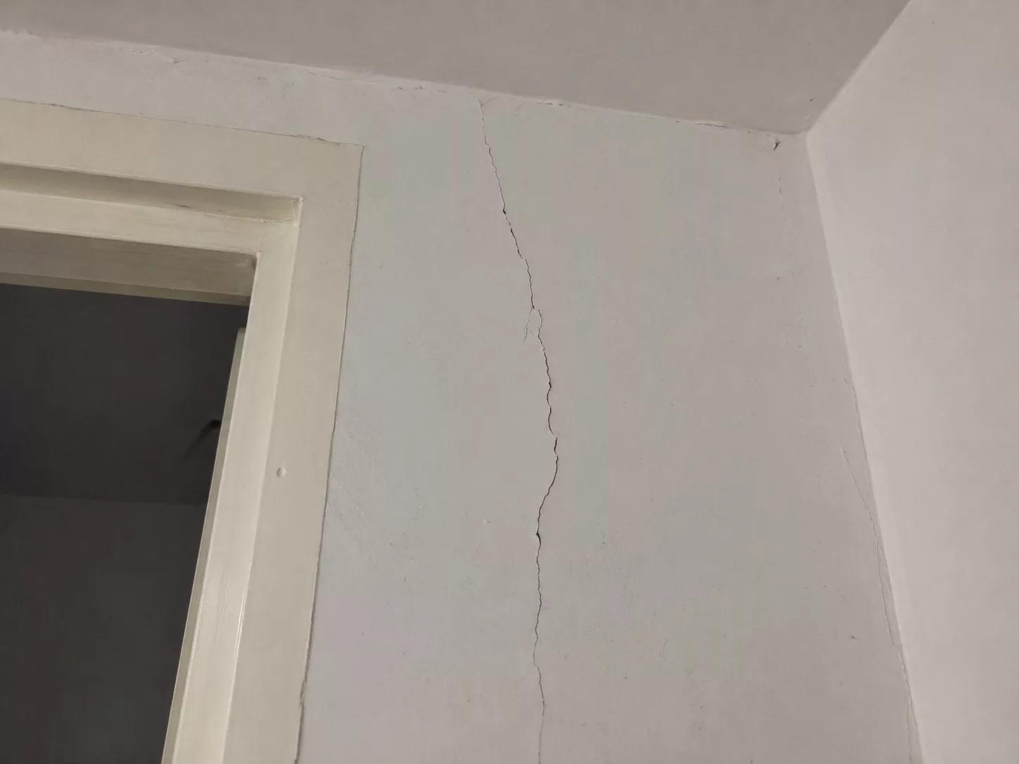 Fissure verticale sur mur en placo dans une maison, près d’un encadrement de porte et du plafond, avec peinture usée et défaut visible.