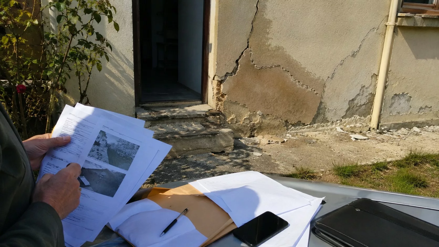Dossier d’assurance et documents de contestation devant une maison fissurée après catastrophe naturelle avec façade abîmée et traces d’humidité