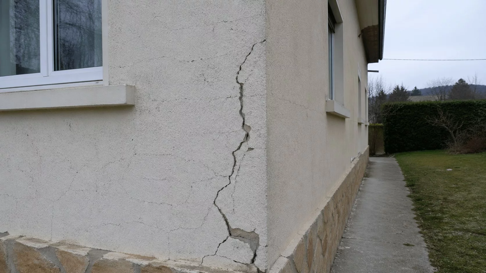 Photo réaliste prise au Panasonic GH4 montrant une fissure verticale importante sur l’enduit d’un mur extérieur de maison, près d’une fenêtre et d’un soubassement en pierre, pour illustrer que faire face à une fissure de maison.