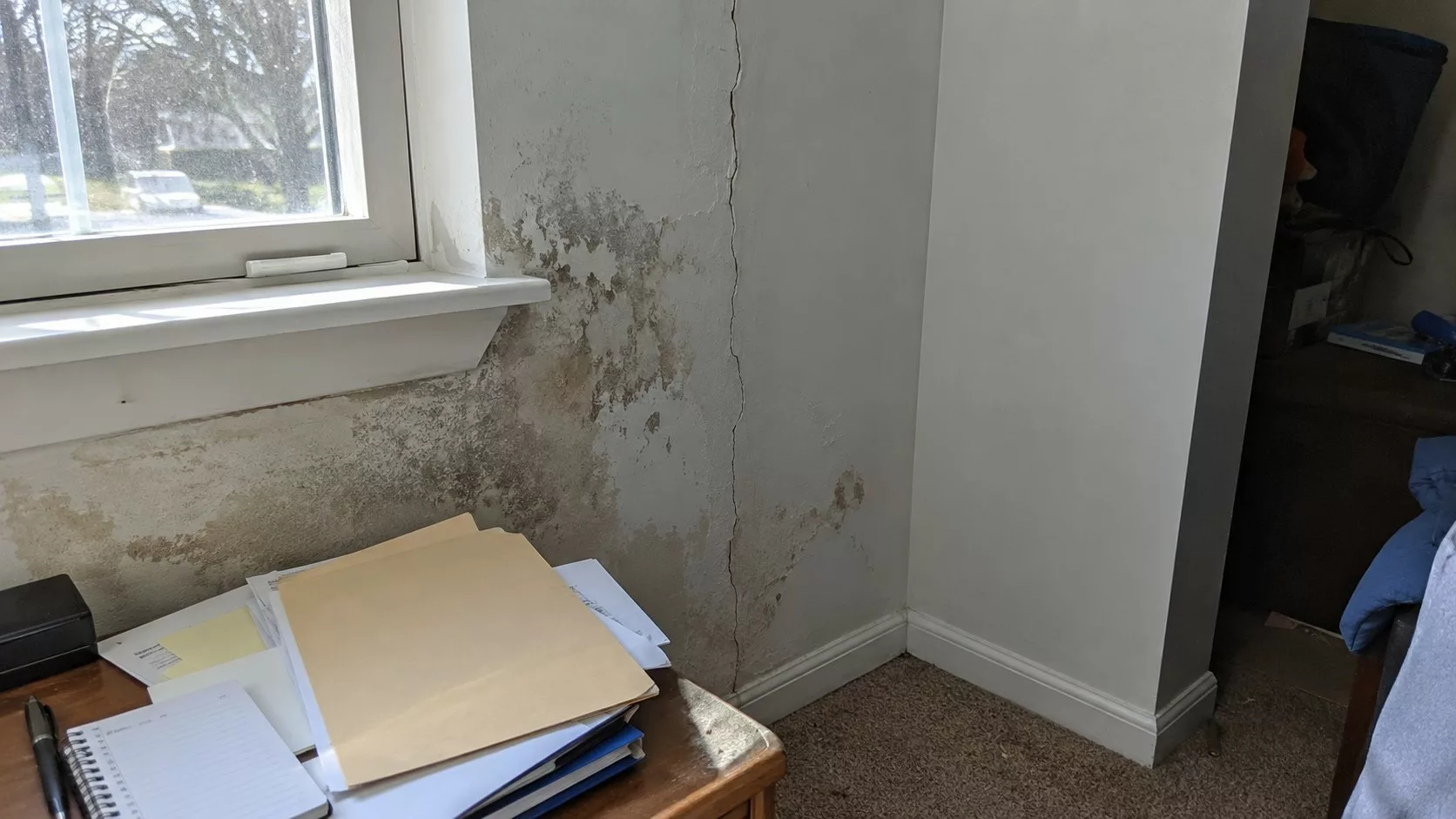 Photo documentaire d’un mur intérieur abîmé près d’une fenêtre, avec fissure verticale, traces d’humidité, peinture écaillée et dossier de documents posé sur une table, illustrant le délai de 2 ans pour agir après la découverte d’un vice caché immobilier.