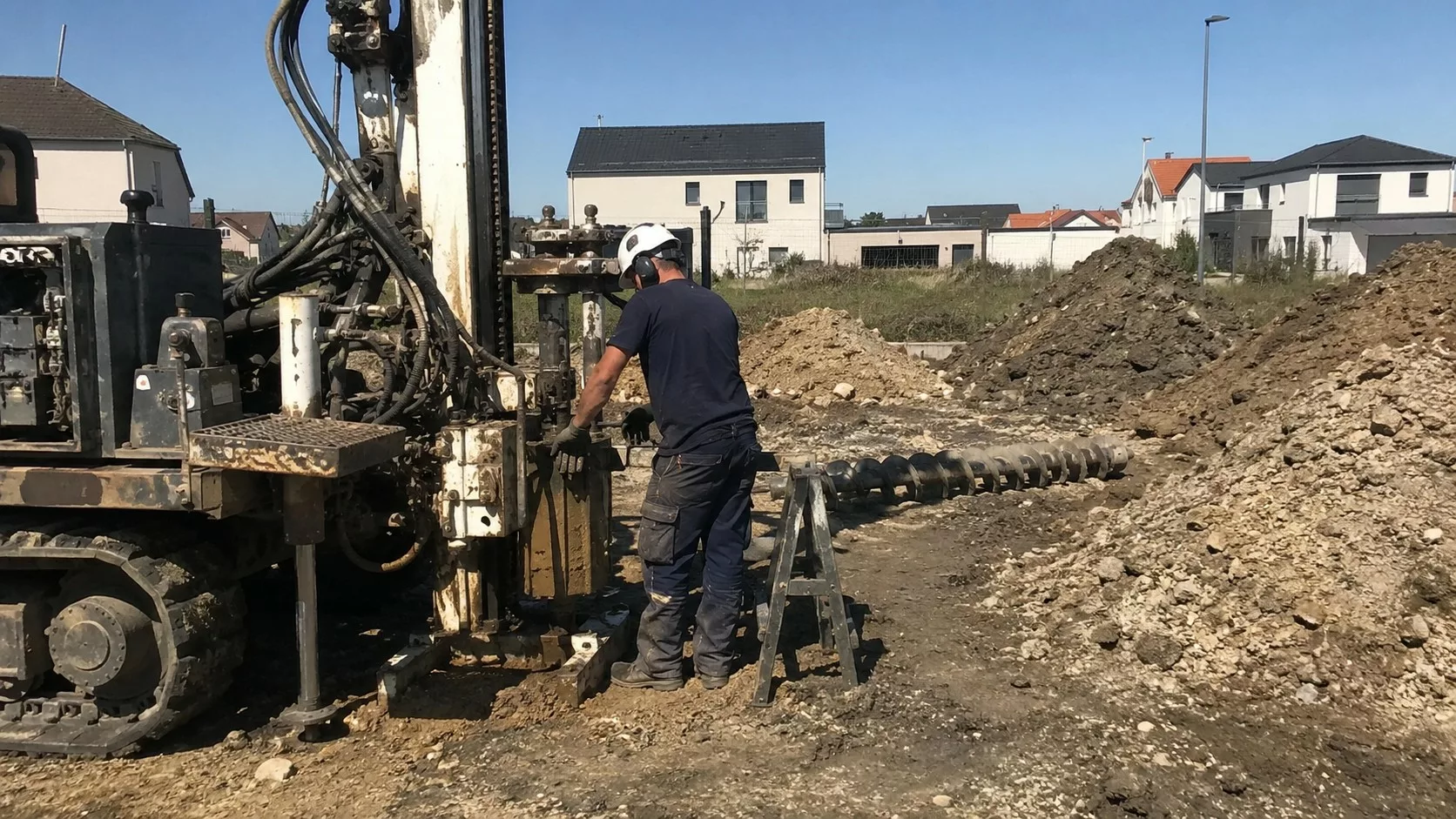 Foreuse de chantier réalisant une étude de sol G1/G2 sur un terrain constructible en plein soleil, avec ouvrier casqué, tas de terre, tarière et maisons en arrière-plan.