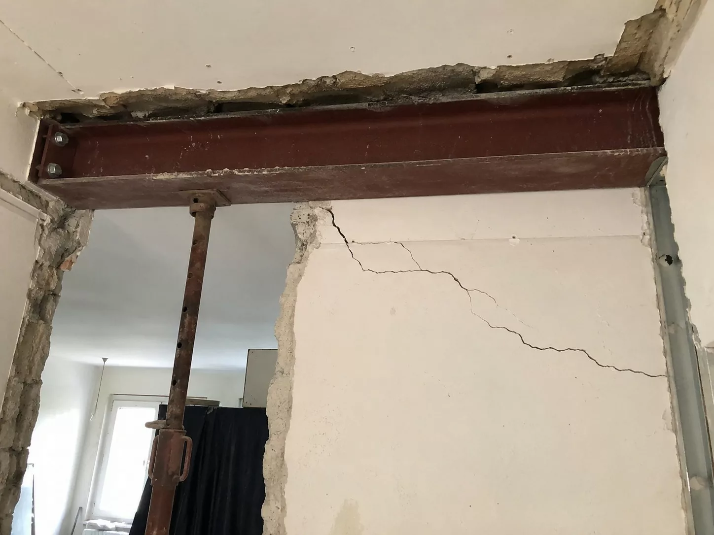 Photo documentaire réaliste format 4/3 montrant des fissures après ouverture d’un mur porteur, avec poutre acier IPN, étai de soutien, mur fissuré, plâtre abîmé et chantier de rénovation intérieur poussiéreux.