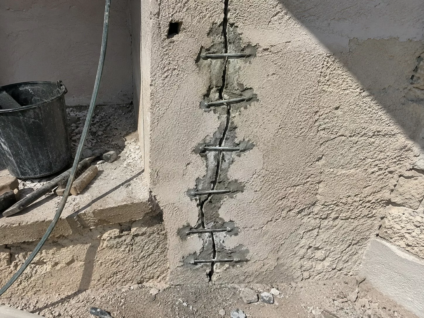 Agrafage d’une fissure verticale sur un mur extérieur avec plusieurs barres métalliques scellées au mortier, seau noir, outils, gravats et chantier en plein soleil, 22 juillet 2026, format 4:3