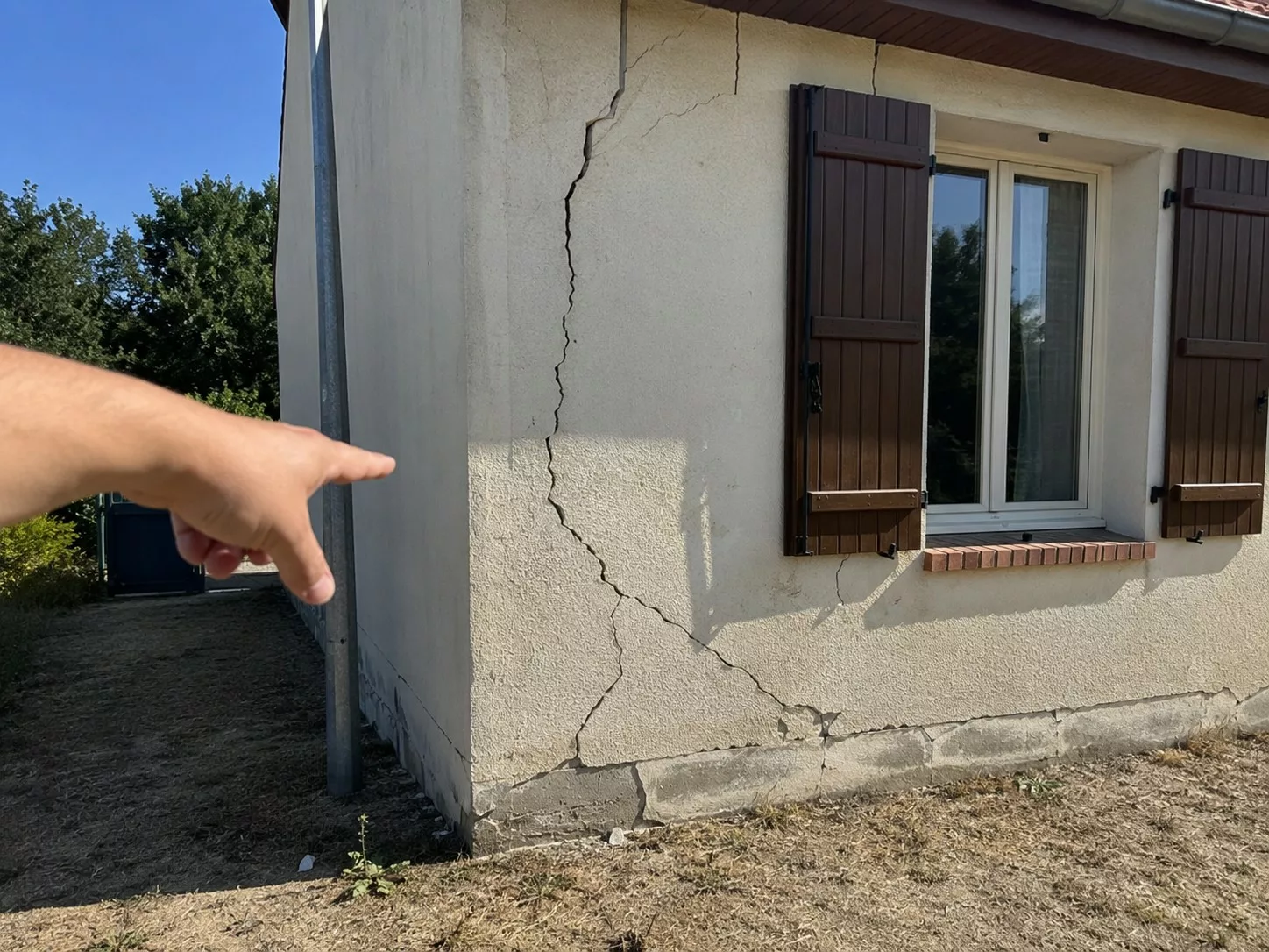 fissures_secheresse_maison_assurance_refus_indemnisation_fissure_verticale_facade_crepie_main_pointant_degats_mur_volets_bois_sol_sec_2026_format_4_3