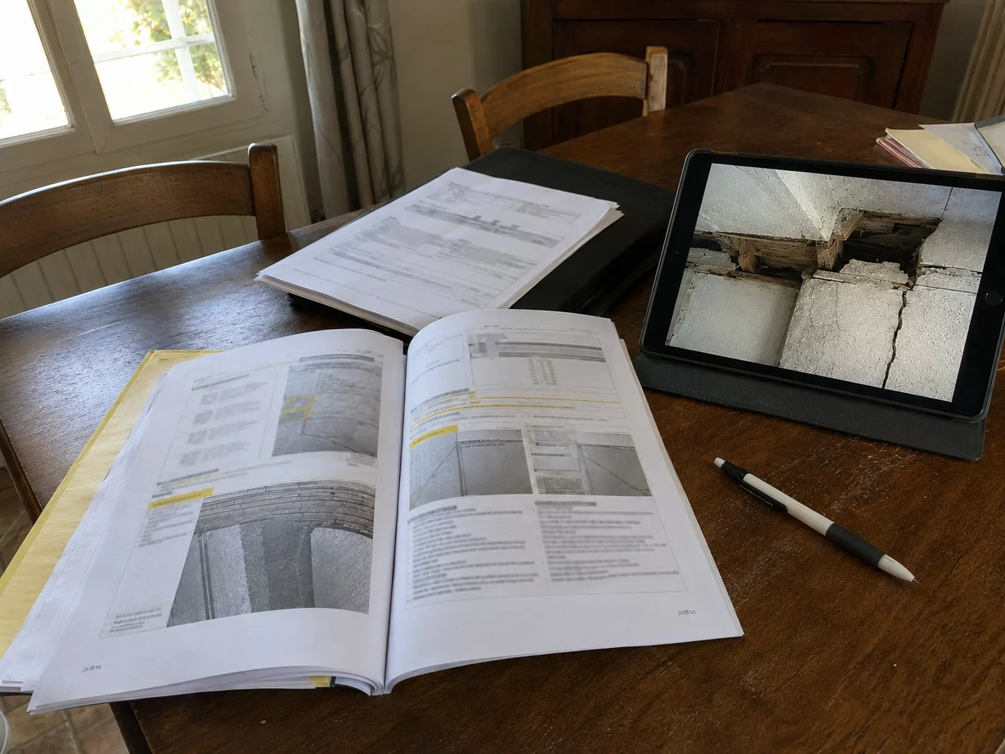 Rapport d'expertise bâtiment consulté sur tablette montrant une fissure structurelle au plafond, documents techniques d'une maison, base d'une négociation avant achat