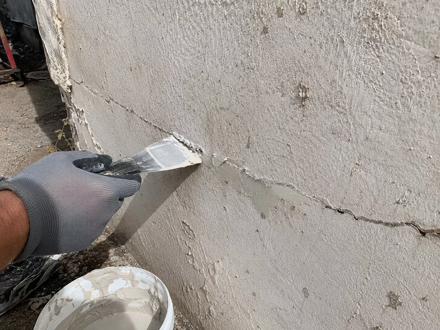 rebouchage d’une longue fissure sur un mur extérieur en enduit avec une spatule, gant de chantier, seau de mortier et application manuelle du produit de réparation, lumière naturelle ensoleillée, 22 juillet 2026, format 4:3