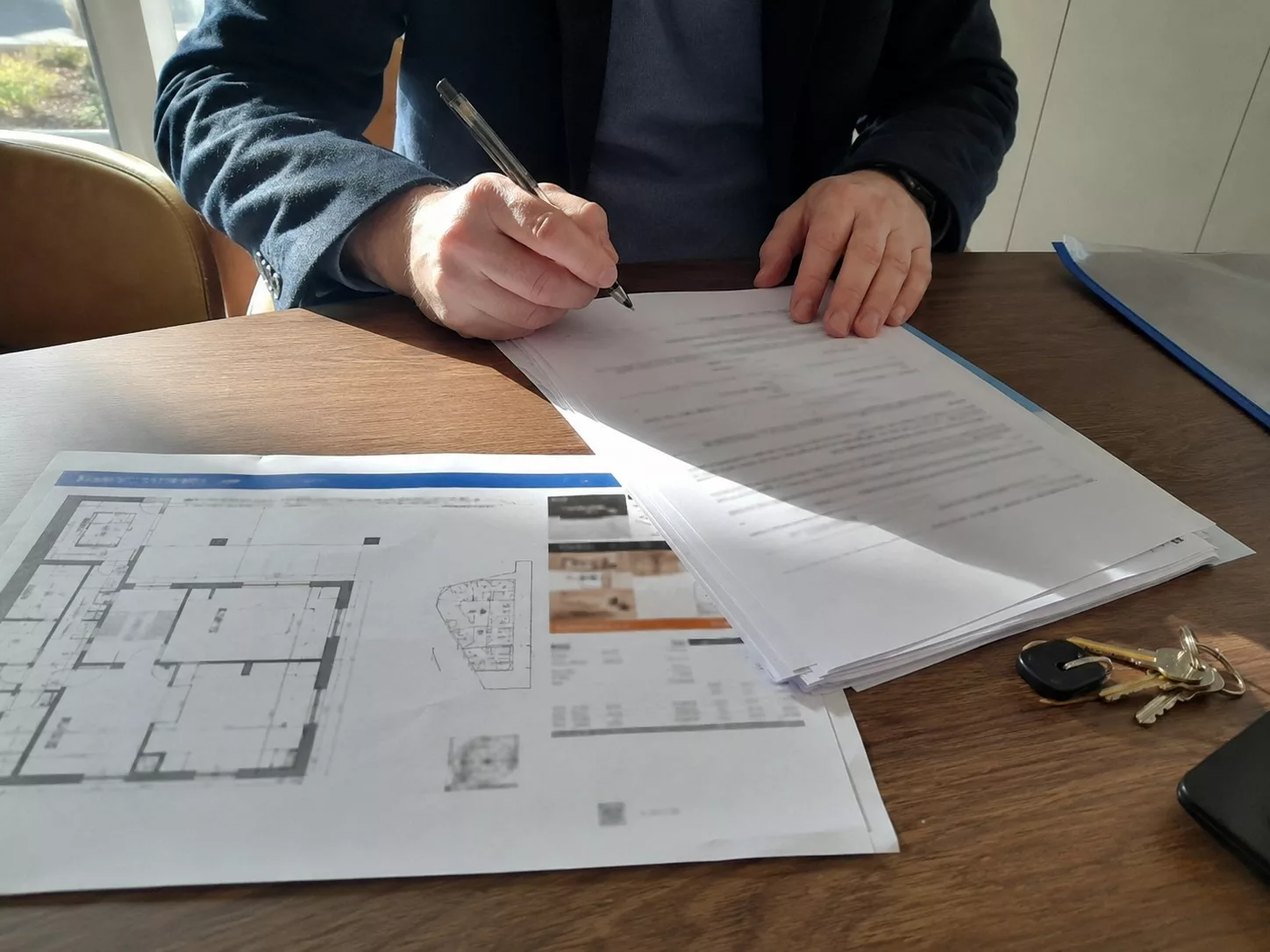 Personne signant un contrat de réservation VEFA sur une table en bois avec plusieurs documents immobiliers, plan détaillé d’un logement neuf, dossier bleu et trousseau de clés, scène réaliste éclairée par une forte lumière naturelle en juillet 2026.
