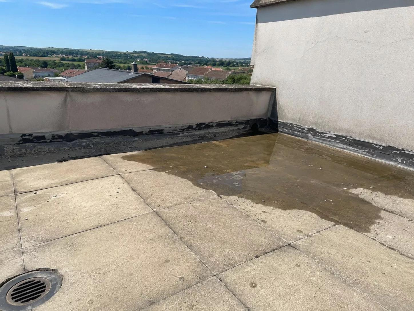 Toiture-terrasse revêtue de dalles grises, large zone d'eau stagnante le long du muret de rive, relevé d'étanchéité noir visible au pied du muret et grille d'évacuation au premier plan