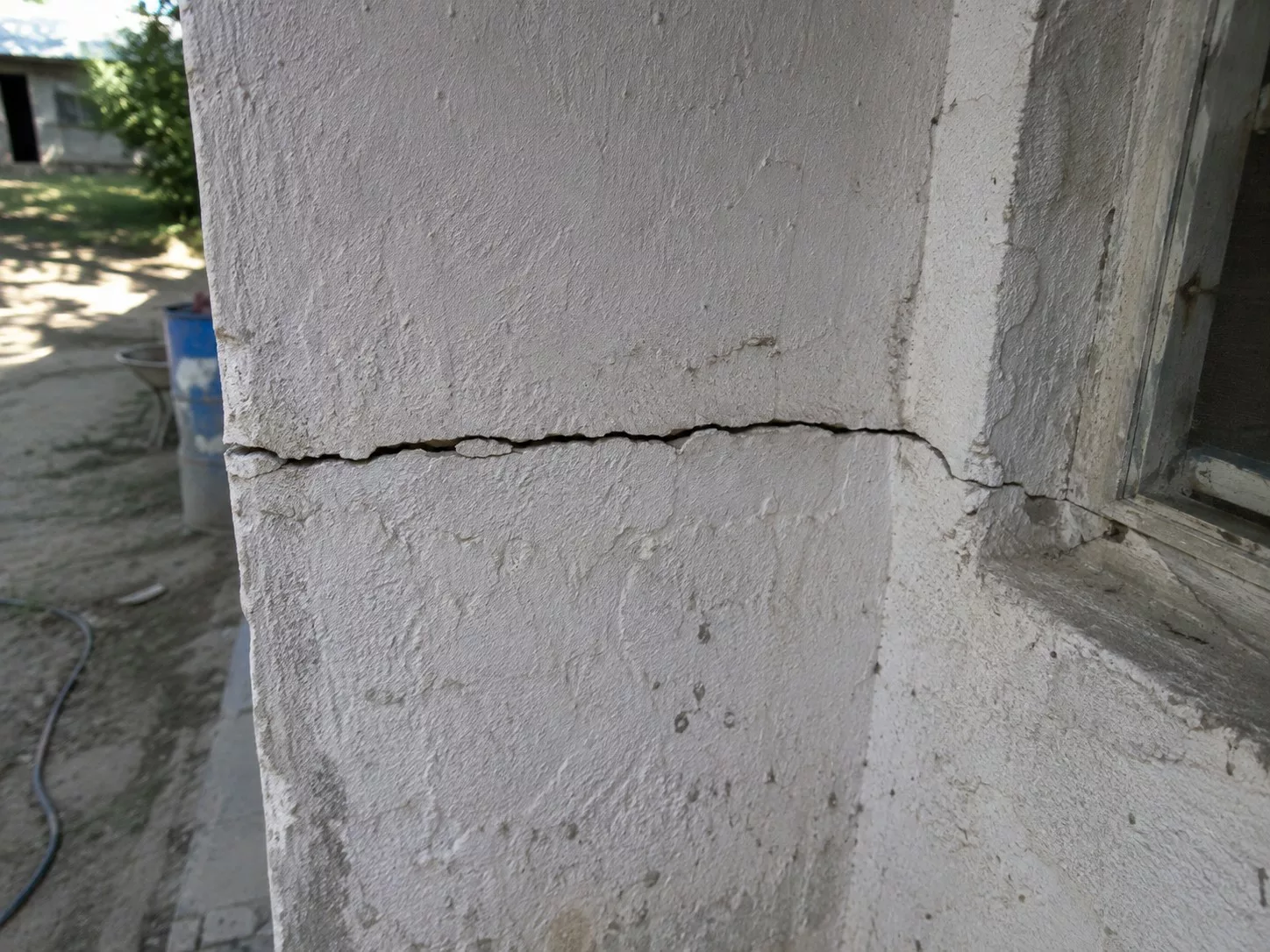 fissure_horizontale_jagee_ouverte_dans_enduit_exterieur_blanc_mur_maconnerie_angle_de_facade_pres_fenetre_crepi_degrade_eclats_salissures_traces_humidite_jardin_ensoleille_arriere_plan_flou_aout_2026