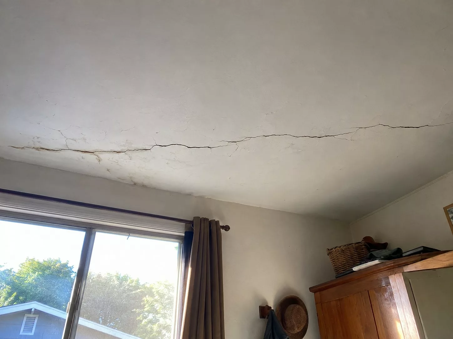 Fissure longitudinale traversant un plafond en enduit peint blanc, microfissures ramifiées, ouverture irrégulière, traces brunâtres d’humidité et décollement localisé près du mur, pièce d’habitation ancienne éclairée par une baie vitrée en plein soleil, armoire en bois et objets du quotidien visibles, prise de vue documentaire grand-angle depuis le sol, perspective ascendante, netteté étendue, lumière naturelle dure, textures de plâtre usé, format 4:3, août 2026
