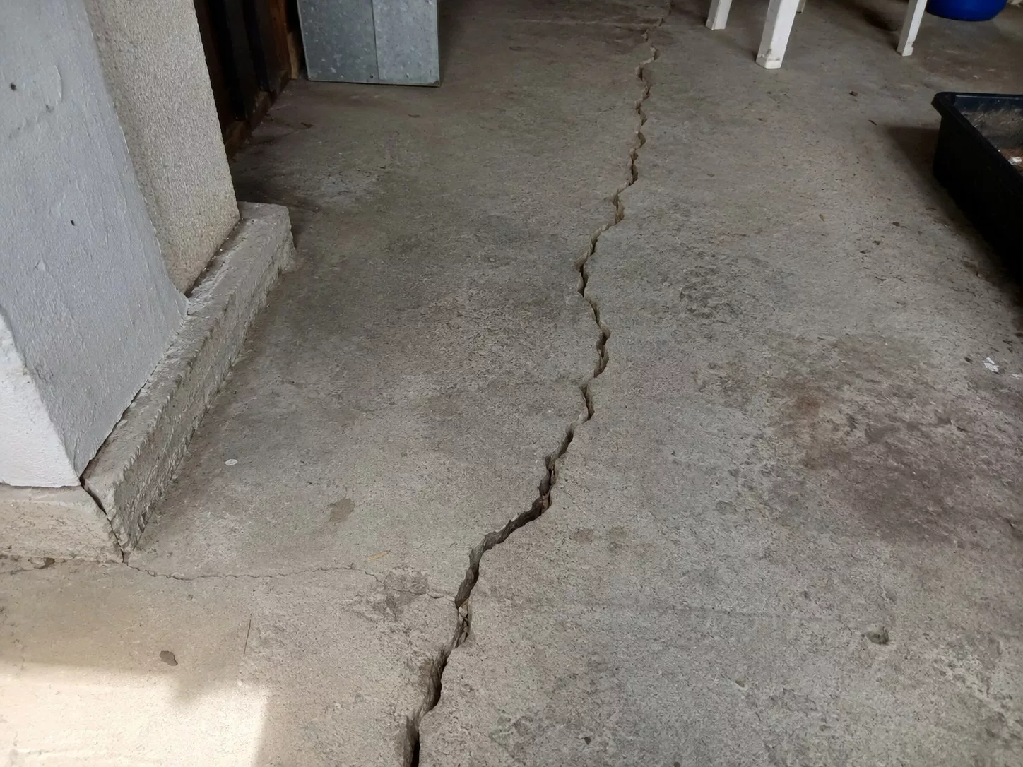 fissure longitudinale irrégulière et profonde traversant un dallage en béton brut dans un garage ou local technique, ouverture visible avec bords écaillés, différence de niveau, poussière, salissures, taches d’usure, petits débris, pied de mur enduit blanc abîmé, bac plastique noir et pieds de chaise en arrière-plan, éclairage naturel ensoleillé, photographie documentaire réaliste au format 4:3, août 2026