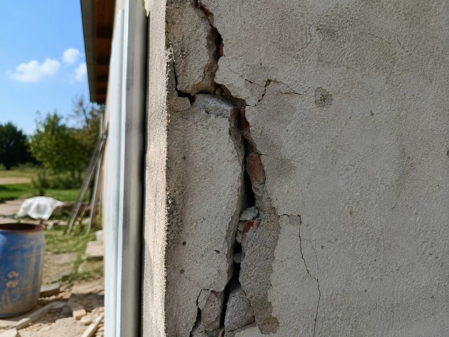 fissure_structurelle_verticale_profonde_sur_mur_porteur_extérieur_enduit_crépi_fendu_et_décollé_maçonnerie_interne_briques_rouges_et_mortier_apparents_lèvres_de_fissure_ouvertes_microfissures_secondaires_débris_en_pied_de_mur_descente_eaux_pluviales_adjacent_arrière_plan_jardin_et_matériel_de_chantier_floutés_lumière_naturelle_ensoleillée_2026