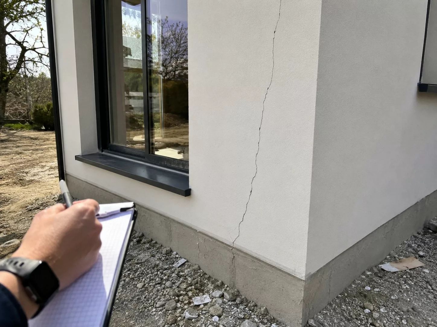 fissure_verticale_longue_sur_enduit_blanc_façade_maison_neuve_angle_de_mur_proche_baie_vitrée_fissure_descendant_jusqu’au_soubassement_béton_main_expert_avec_stylo_et_bloc-notes_inspection_sur_site_sol_gravillonné_lumière_naturelle_ensoleillée_17-08-2026