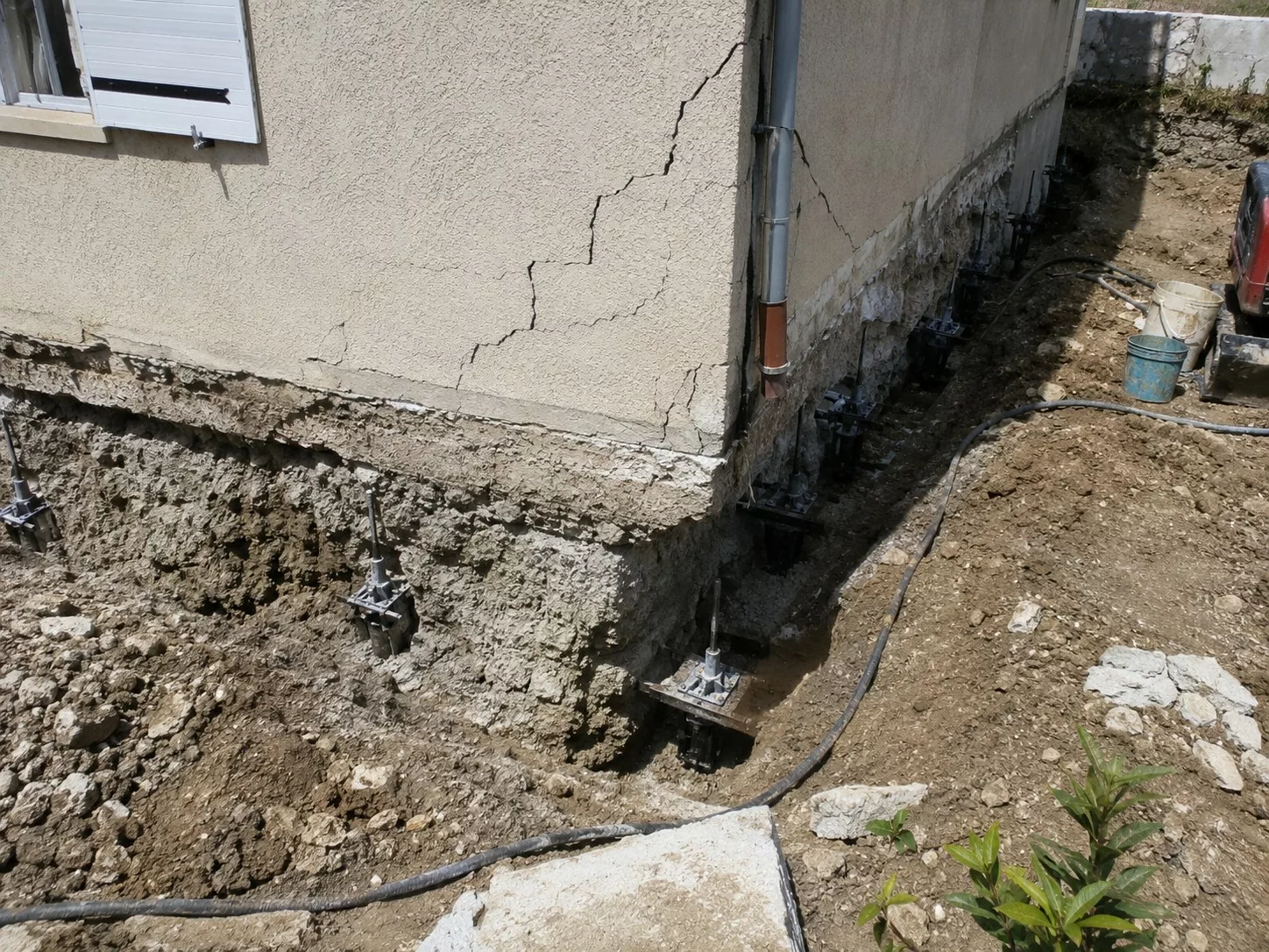 Micropieux sur maison fissurée en 2026, façade en enduit présentant plusieurs fissures structurelles, fondations mises à nu par excavation périphérique, reprise en sous-œuvre avec têtes de micropieux métalliques et platines de renforcement sous les murs porteurs, tranchée de chantier, sol remanié, gravats, flexibles et matériel de fondation, intervention de stabilisation d’une maison affectée par des mouvements de terrain.