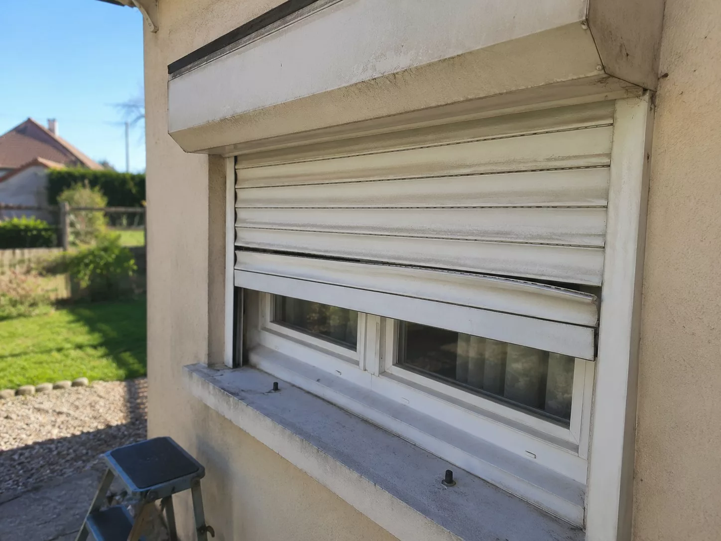 Volet roulant électrique bloqué à mi-hauteur sur la fenêtre d'une maison, lames blanches désalignées et lame inférieure tordue, coffre de mécanisme poussiéreux, escabeau posé sous le volet