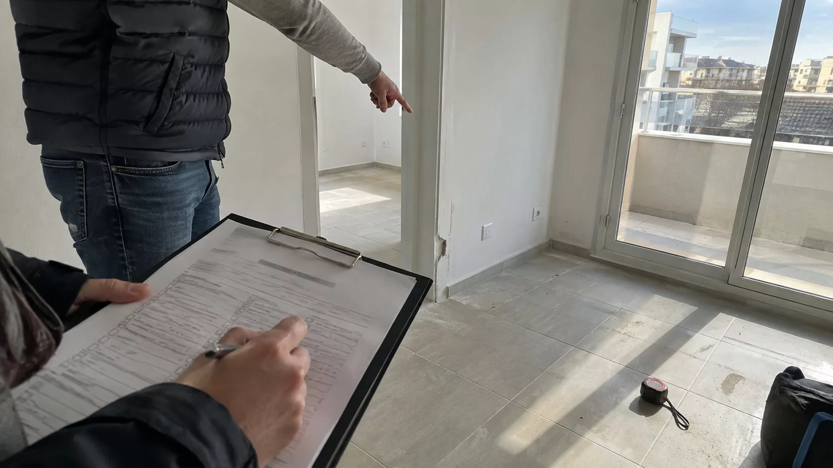Procès-verbal de livraison VEFA sur porte-documents au premier plan, professionnel pointant une réserve sur un angle de mur écaillé dans un appartement neuf lumineux avec baie vitrée et mètre ruban au sol
