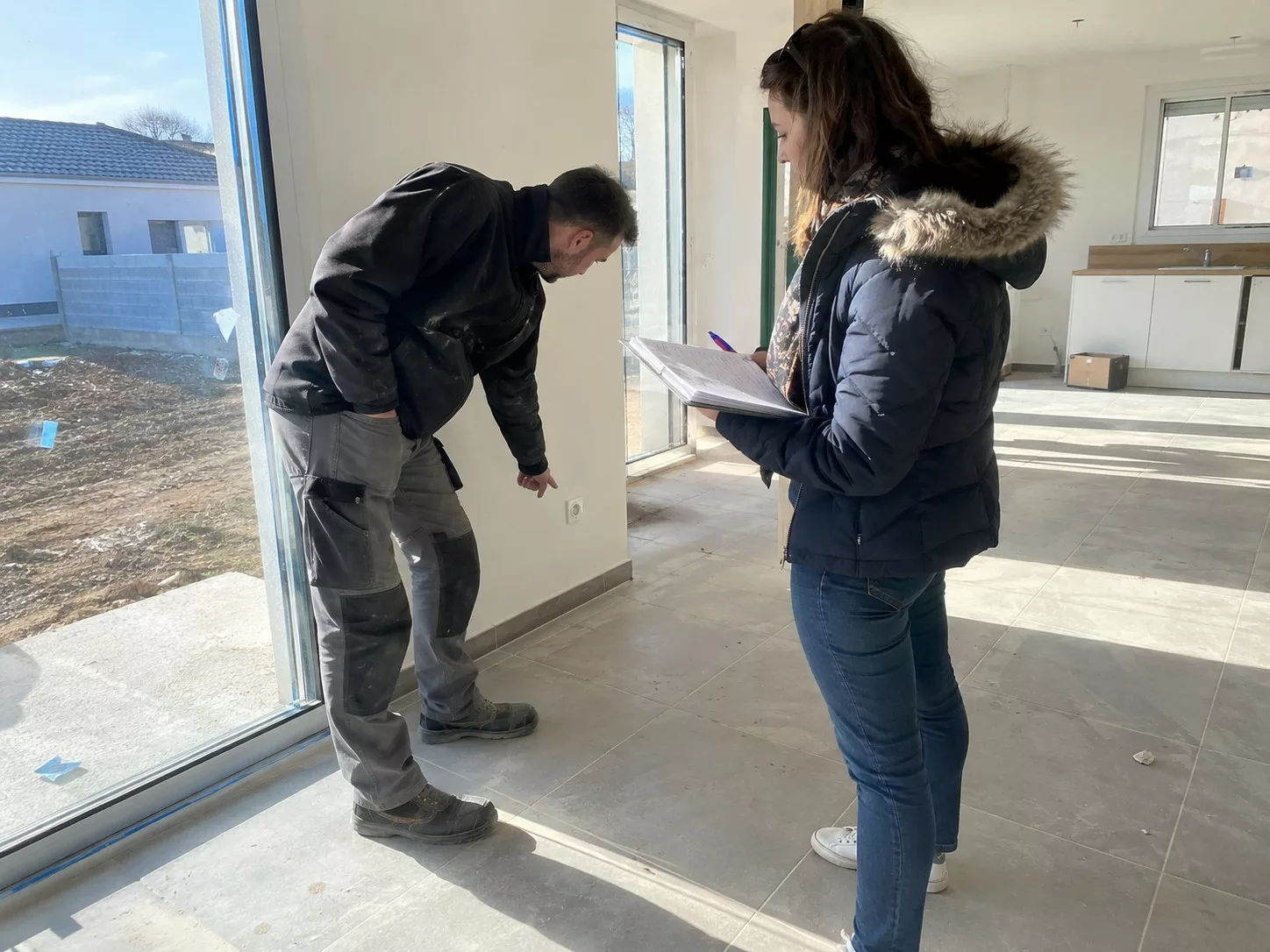 Réception d'une maison neuve, professionnel du bâtiment contrôlant une prise près d'une baie vitrée, propriétaire tenant une check-list et un stylo