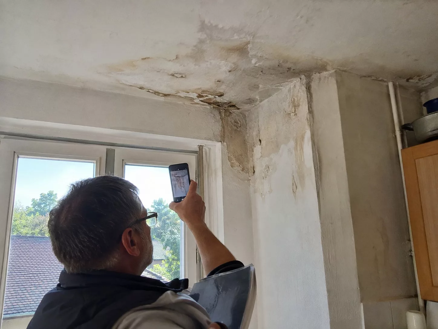 Homme photographiant au smartphone une infiltration au plafond, dans l'angle au-dessus d'une fenêtre, enduit taché et dégradé sur le plafond et le mur